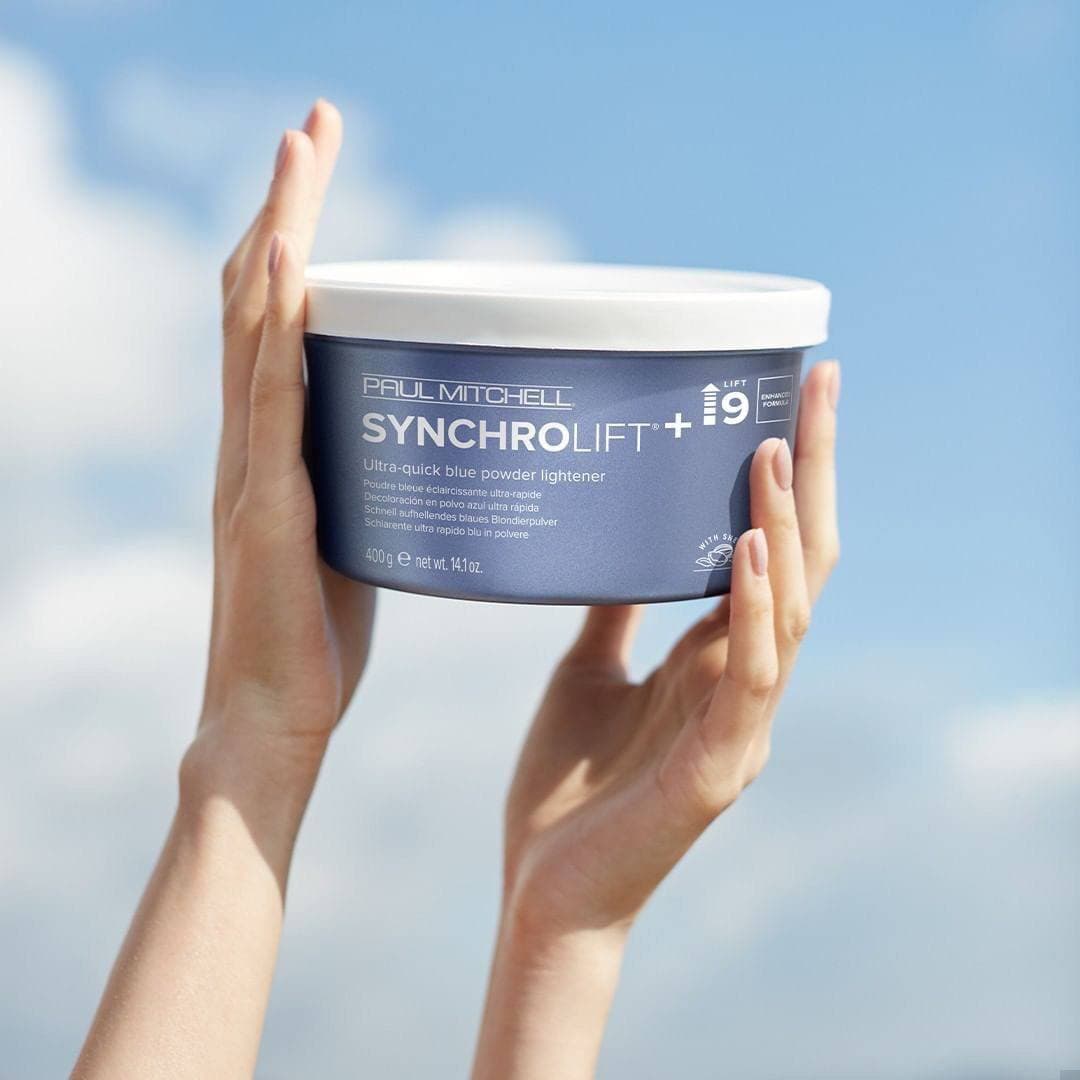 Руки держат банку осветляющей пудры Paul Mitchell SynchroLift+9 на фоне голубого неба